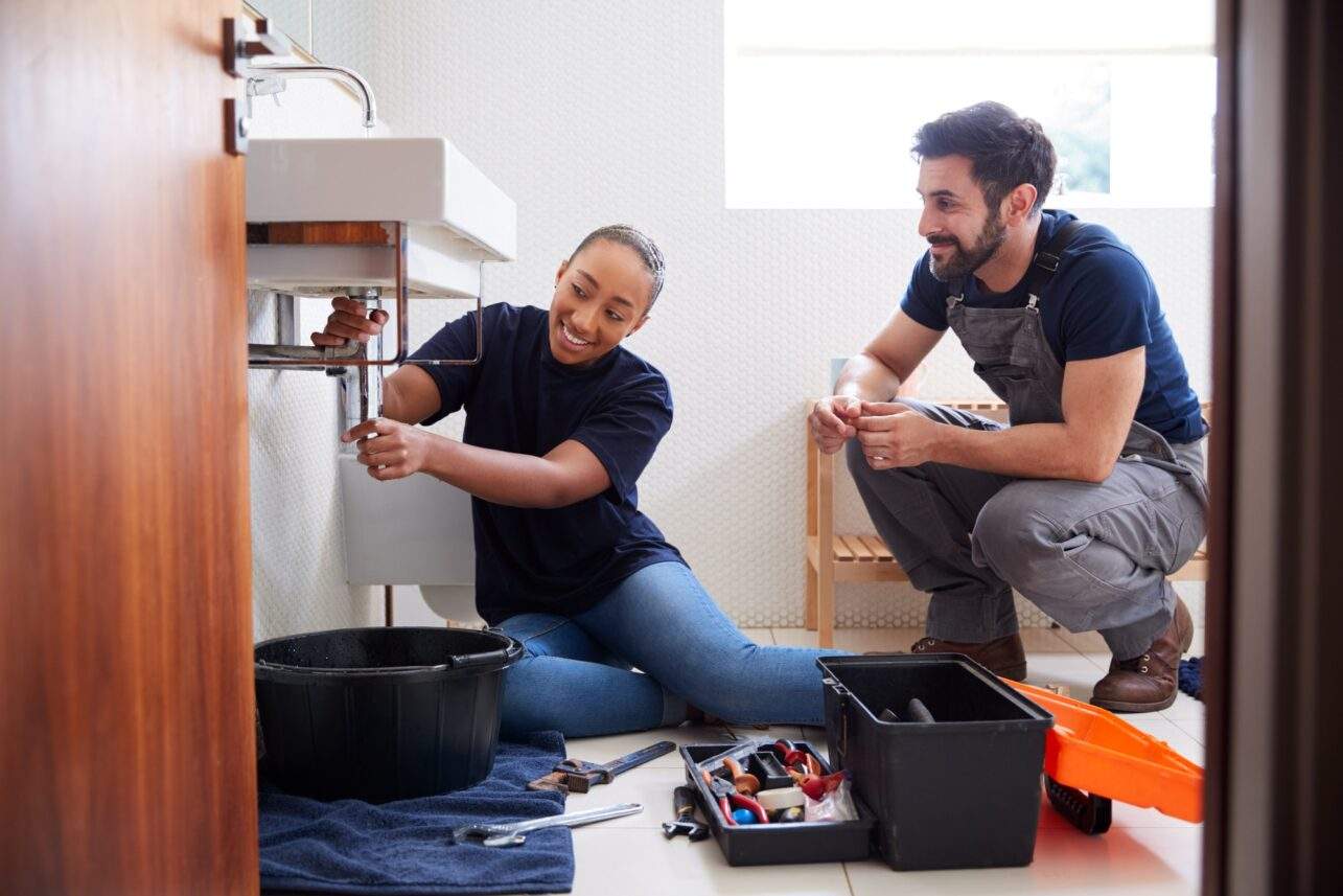 Male Plumber Teaching Female Apprentice To Fix Leaking Sink In Home Bathroom